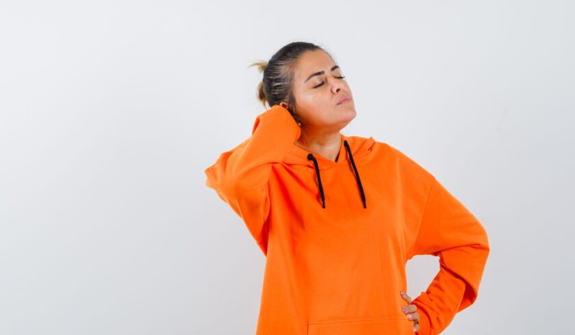 woman in orange hoodie keeping hand behind head and looking relaxed , front view.